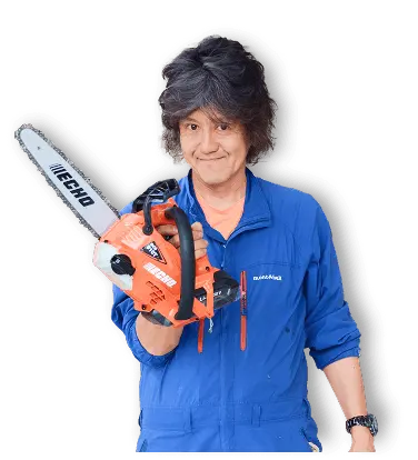 田中機械社長（タナカヨシミ）が青いmont-bell作業服上下を着用し、チェーンソーを片手で持って笑顔でポーズを取っている