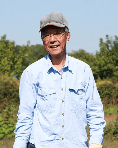 笑顔で立つ高齢男性農業従事者、水色のワークシャツと帽子を着用し、緑豊かな農園を背景に