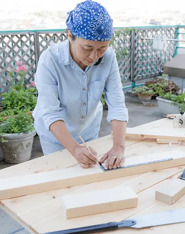 青いバンダナを着用した職人が木材を加工している様子、屋外の作業台で丁寧に作業中